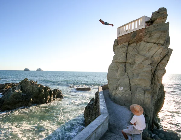 A cliff diver mid-air after jumping from a platform on a rocky outcrop in Mazatlán, Mexico, with the ocean in the background.
