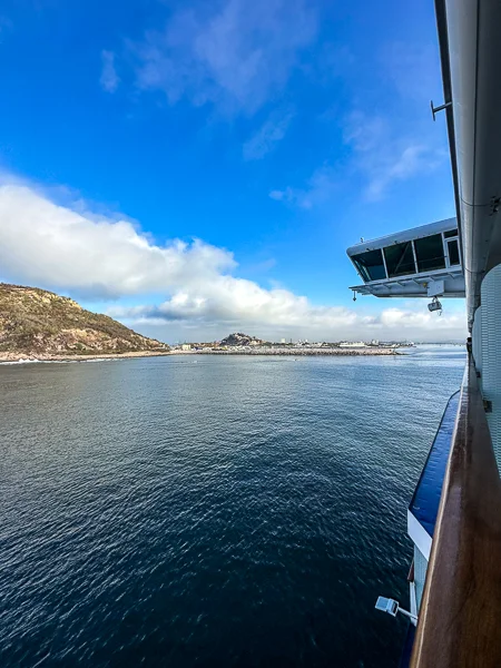 View of Mazatlán from a cruise ship approaching the port, with the coastline and hills in the distance under a blue sky.