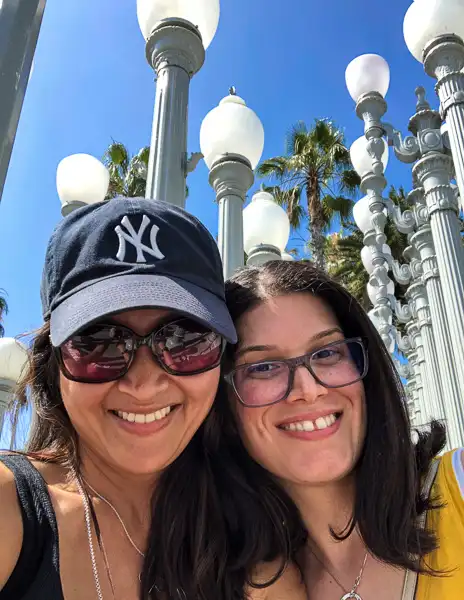 kathy and best friend take a selfie in front of the iconic Urban Lights installation at LACMA, surrounded by rows of vintage street lamps against a backdrop of palm trees and clear blue skies.