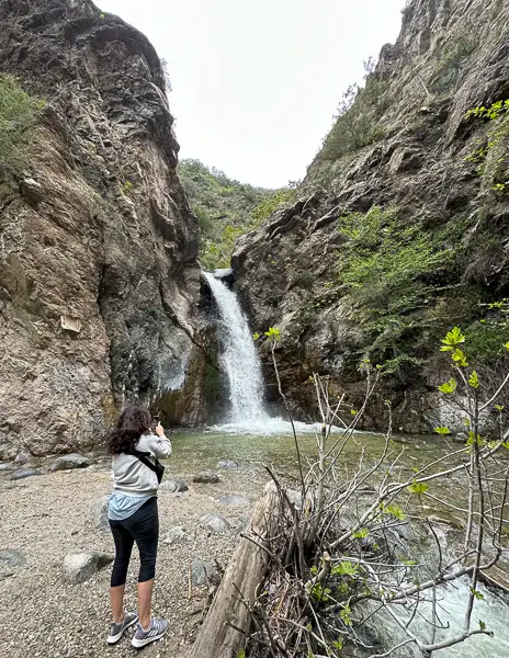 A person standing at the base of Eaton Canyon Waterfall in Pasadena, capturing a photo of the waterfall as it cascades down the rocky cliffs into a small pool below.