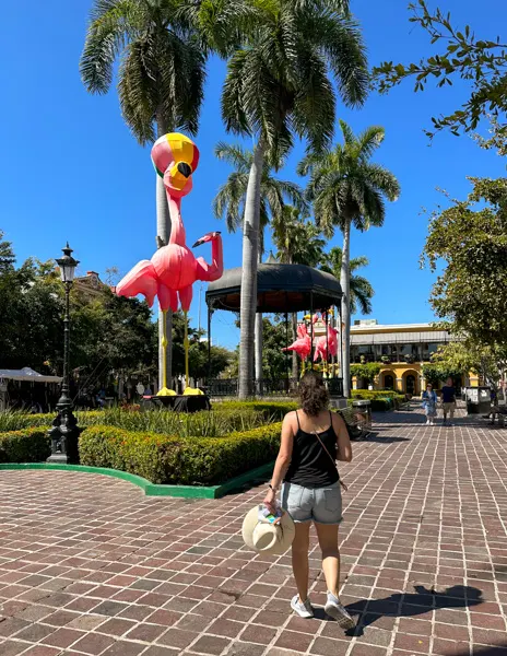 A woman (kathy) walking through Plaza Machado in Mazatlán, surrounded by palm trees and large pink flamingo statues