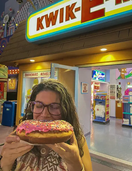 woman biting into a pink lards donut in front of kwik e mart at universal studios hollywood
