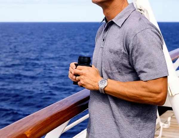 featured blog image | cruise wear for men | man in blue polo shit, holding binoculars standing by a cruise railing