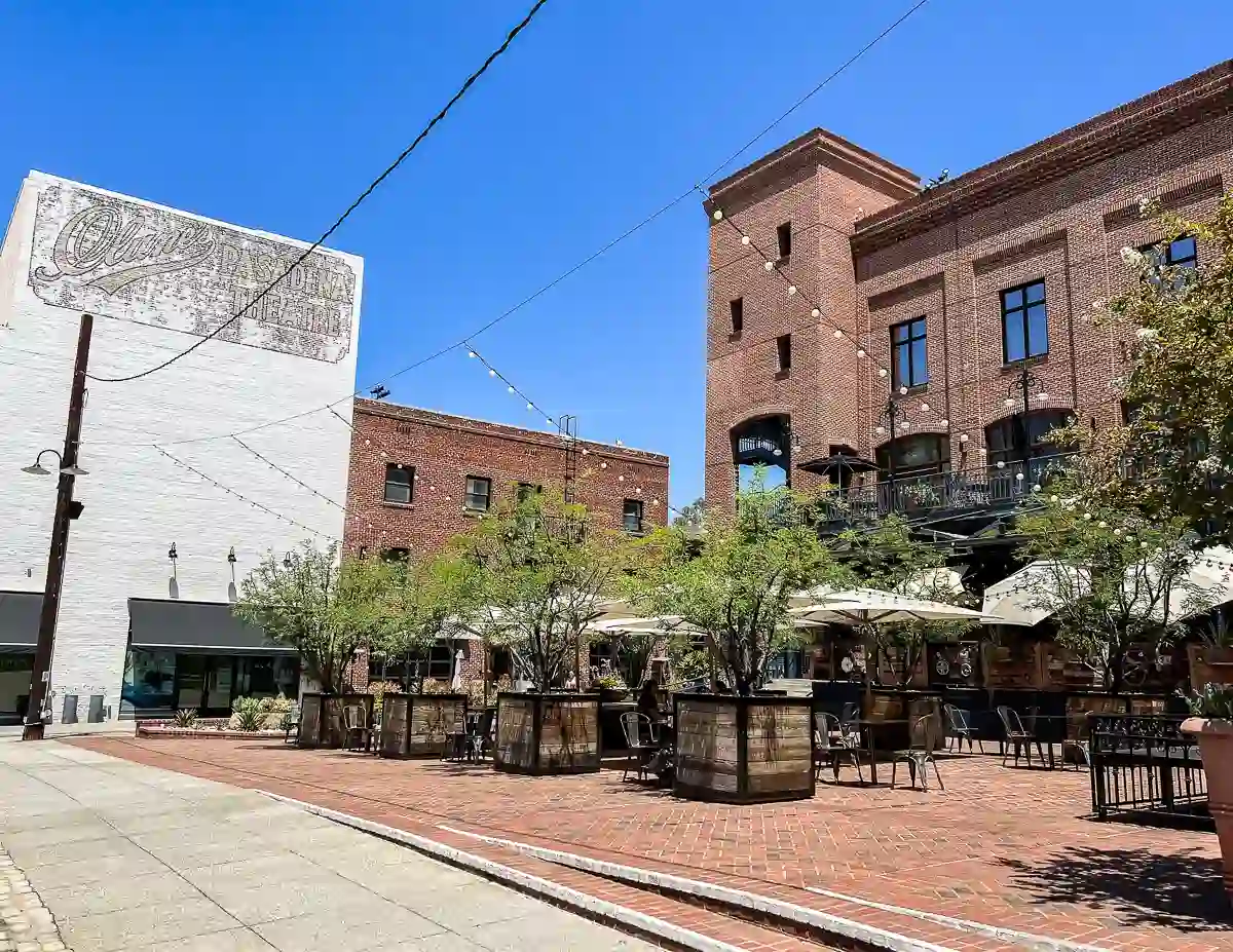 Open-air patio with tables under umbrellas in Old Pasadena, surrounded by historic brick buildings and a vintage advertisement on the wall.