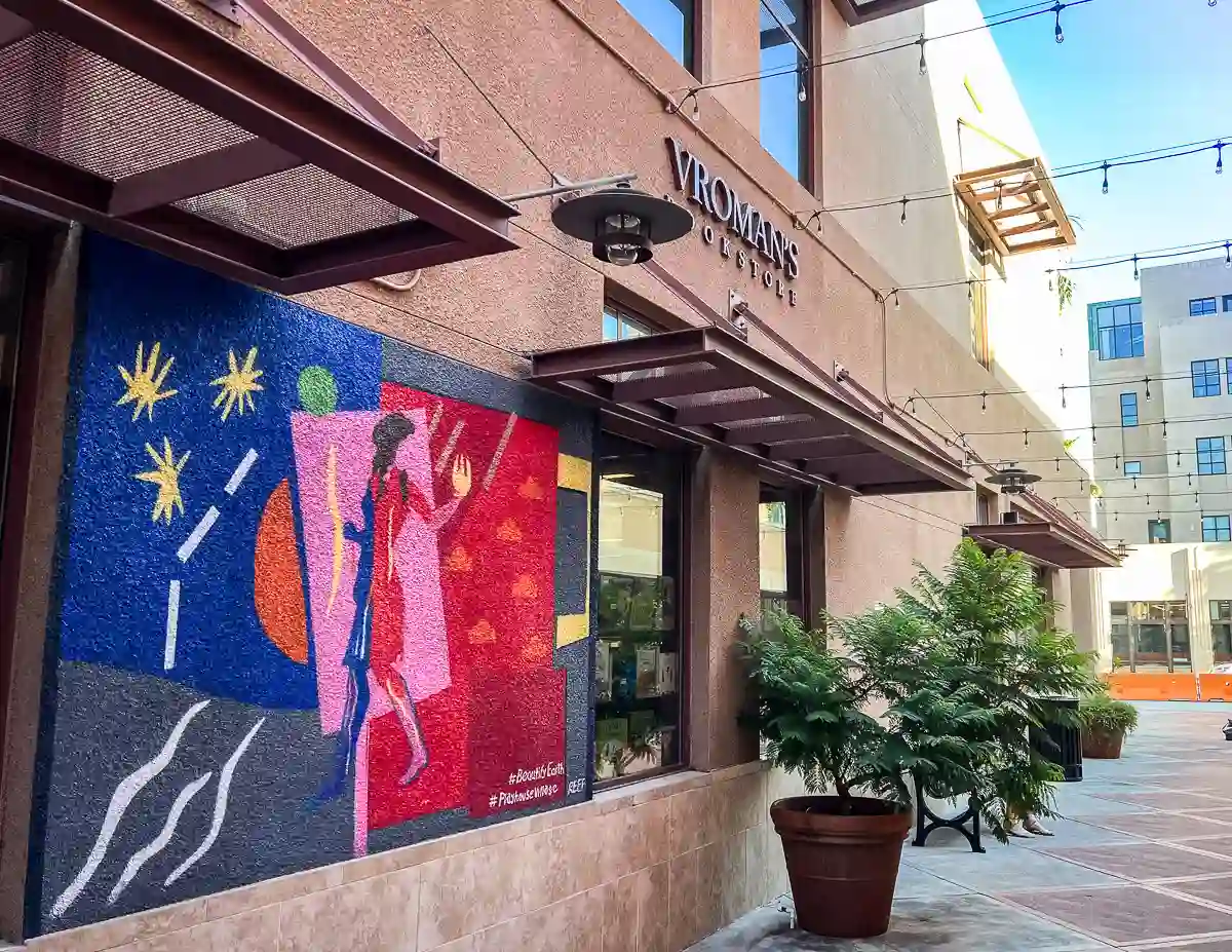 Side view of Vroman's Bookstore in Pasadena, featuring a colorful mural on the wall with abstract shapes and a figure walking, illuminated by string lights in the alleyway.