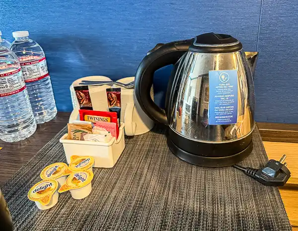 Electric kettle and tea setup in a cruise cabin, with tea bags, coffee packets, creamers, and bottled water arranged on the counter.