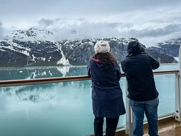 Cruise passengers (Kathy and her husband) on a balcony watching glaciers and snow-covered mountains during an Alaska cruise