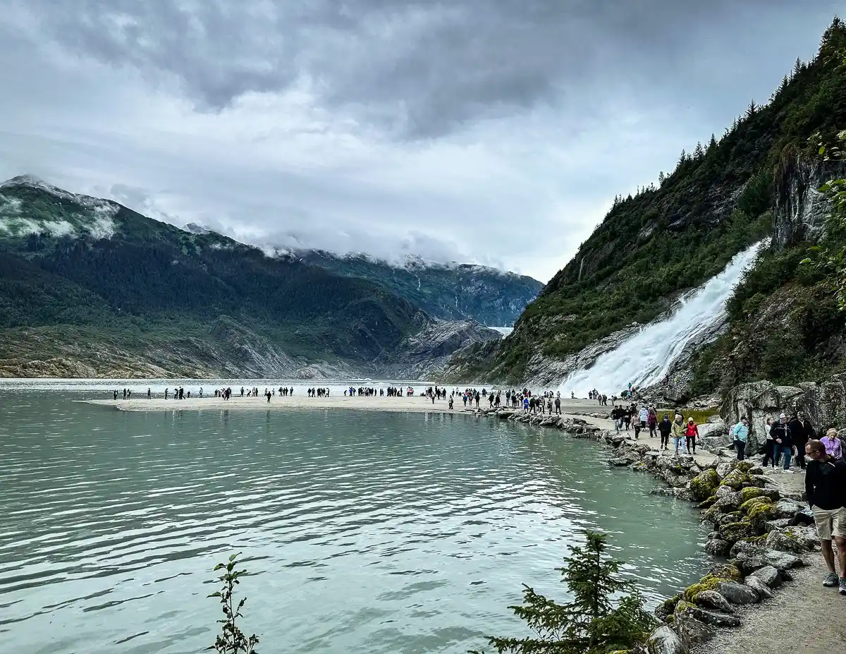 Visitors walking along a scenic trail near Nugget Falls in Juneau, Alaska, with the waterfall cascading down a rocky mountainside and lush greenery surrounding the area. The cloudy sky and mist add to the dramatic natural landscape.