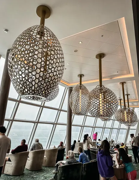 The main observation lounge on the Norwegian Encore, featuring large floor-to-ceiling windows, unique decorative lighting, and guests enjoying the view.