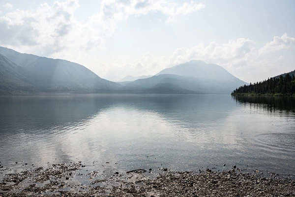 "A serene view of Tutshi Lake with a pebbly shoreline in the foreground, calm reflective waters, and surrounding mountains partially obscured by hazy skies, capturing a tranquil and scenic Alaskan landscape.