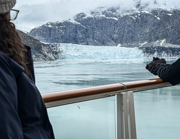 Two cruise passengers (kathy and her husband) standing at the ship’s railing admiring Margerie Glacier in Glacier Bay, Alaska, with calm icy water and rugged snow-dusted mountains in the background.