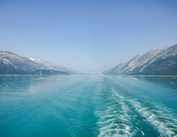 
A tranquil view of Glacier Bay from the aft of a cruise ship, showcasing the ship's wake cutting through the turquoise waters, with scattered ice floating and snow-capped mountains in the distance under a clear blue sky.