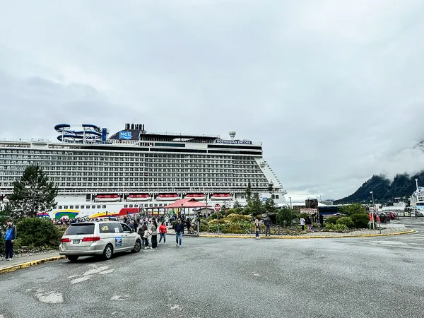 the Norwegian Encore cruise ship docked at Juneau, Alaska. The massive ship is docked beside a busy terminal in Juneau with pedestrians and vehicles moving about, set against a backdrop of low-hanging clouds and distant mountains.
