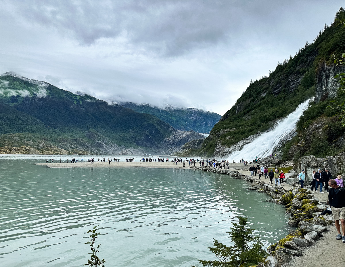 Nugget Falls with visitors walking along the shore in Mendenhall Glacier National Park, showcasing the waterfall's impressive scale