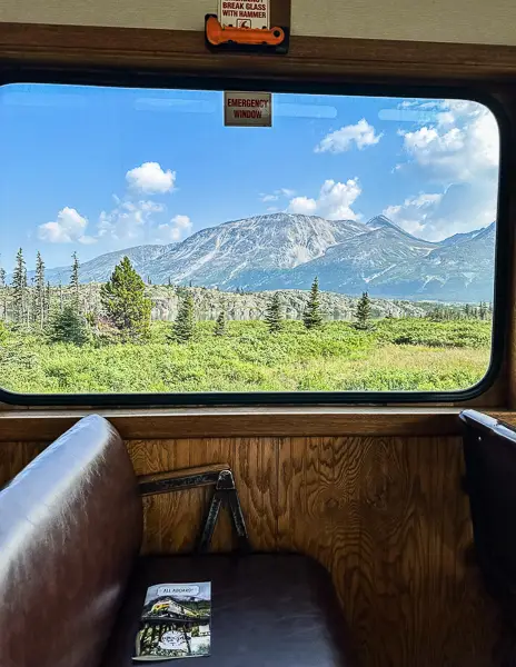 View from inside a vintage train on the White Pass Railway, looking out an emergency window at a stunning Alaskan landscape featuring a vast, tree-dotted plain leading up to a prominent mountain under a clear blue sky.