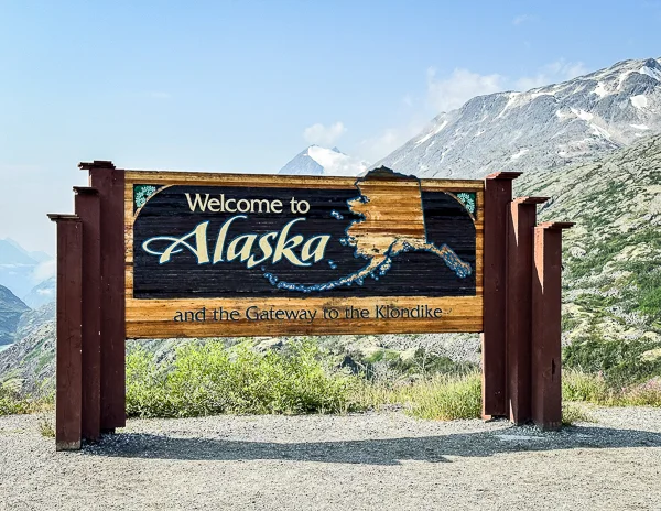 The 'Welcome to Alaska and the Gateway to the Klondike' sign, featuring a large wooden signboard set against a backdrop of mountains and blue sky, marking a significant tourist spot in Alaska.