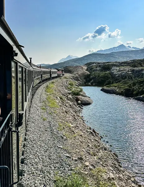 View from a moving train showing another train traveling along a narrow track beside a calm body of water, surrounded by rugged terrain under a clear blue sky.