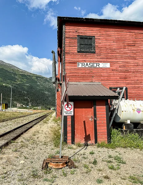 A view of the rustic red Fraser train station against a backdrop of green mountains, clear skies, and adjacent railway tracks.