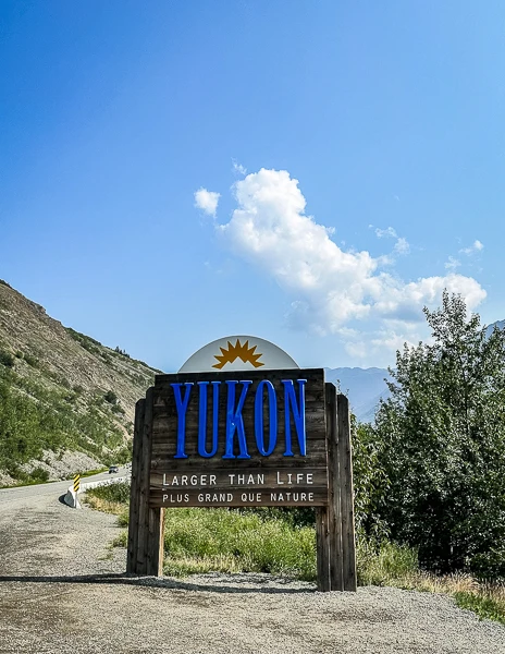 A rustic wooden sign reading 'YUKON - Larger Than Life, Plus Grand Que Nature' at the entrance of Yukon Territory, with a mountainous landscape in the background under a clear blue sky.