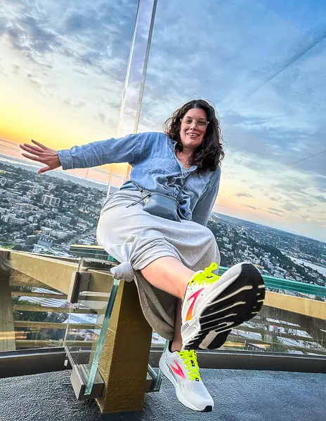 kathy from tasty itinerar leaning up against the glass of the seattle's space needle titling glass with city views of Seattle at sunrise
