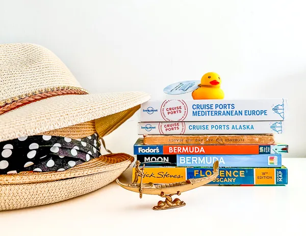 yellow cruising duck sitting on a pile of travel guide books, a bronze venetian gondola in front of it and a pile of hats next to it