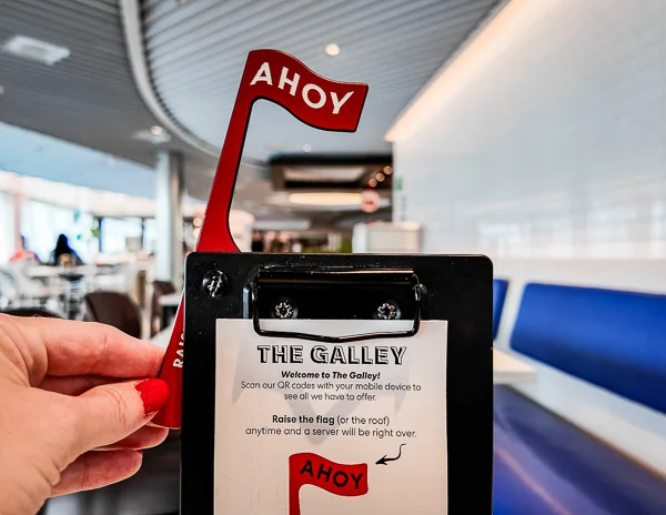 A hand is turning up the red “AHOY” service flag at The Galley on Virgin Voyages, indicating the call for a server, with the dining area visible in the background.