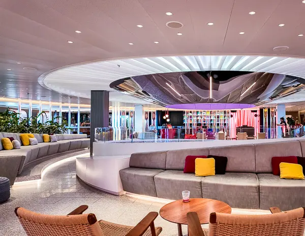 a contemporary lounge with a curved grey sofa, colorful pillows, wooden chairs, and round tables along the roundabout of scarlet lady atrium