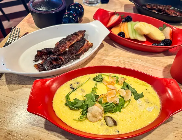 A brunch spread on a table featuring a dish featuring a baked frittata topped with greens and mushrooms, a side plate of crispy bacon, and a red bowl of fresh fruit including melon slices, berries, and pineapple.