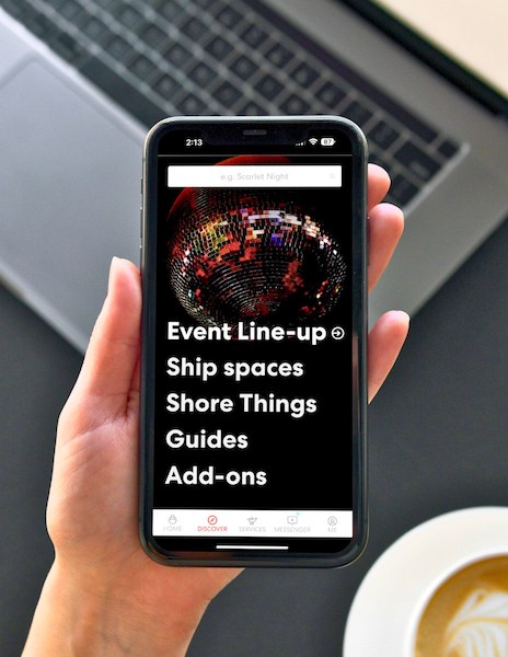 A hand holding a smartphone with the Virgin Voyages app open, displaying options like 'Event Line-up', 'Ship spaces', 'Shore Things', 'Guides', and 'Add-ons', with a laptop and a cup of coffee in the background.