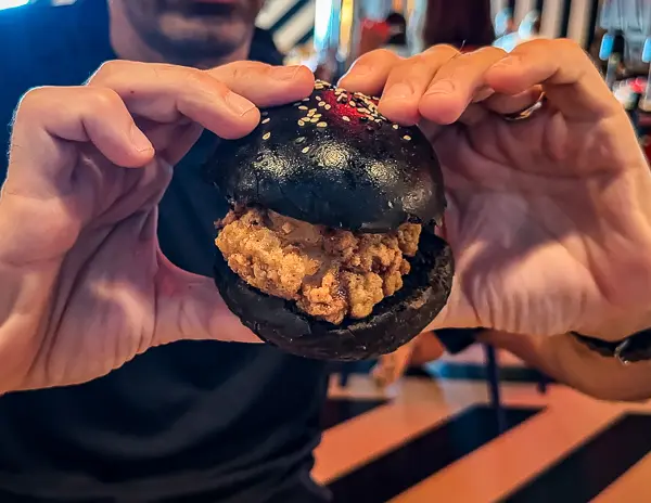 A person holding up a black bun fried chicken sandwich, showcasing the crispy chicken and seed-topped bun, on Virgin Voyages.