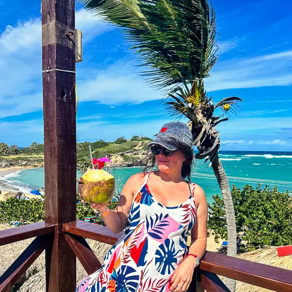 kathy holding a coconut with an umbrella and straw with a beautiful beach behind her in the dominican republic