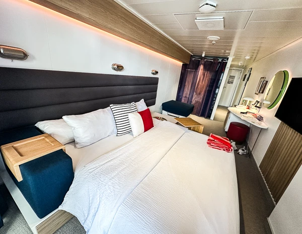 Virgin Voyages cruise ship cabin featuring a sofabed with white bedding, red and striped accent pillows, a side table, a desk area, and a closed curtain with a hint of the ocean view beyond.