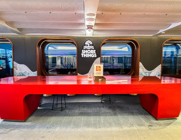 Inside the Scarlet Lady ship by Virgin Voyages, a vibrant red desk labeled "SHORE THINGS" for excursion planning and booking, with large windows overlooking the ocean in the background.