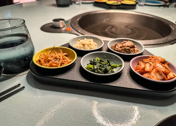 An assortment of small Korean BBQ bites, including kimchi and seasoned vegetables, arranged around a circular cooking station on Virgin Voyages