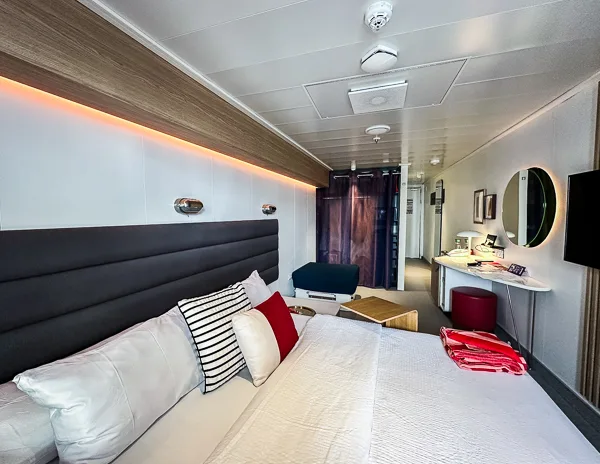 Scarlet Lady xl jūros terasos kajutė su didele lova su minkšta patalyne, svetainės zona su sofa, akcentinėmis pagalvėmis ir kosmetine.