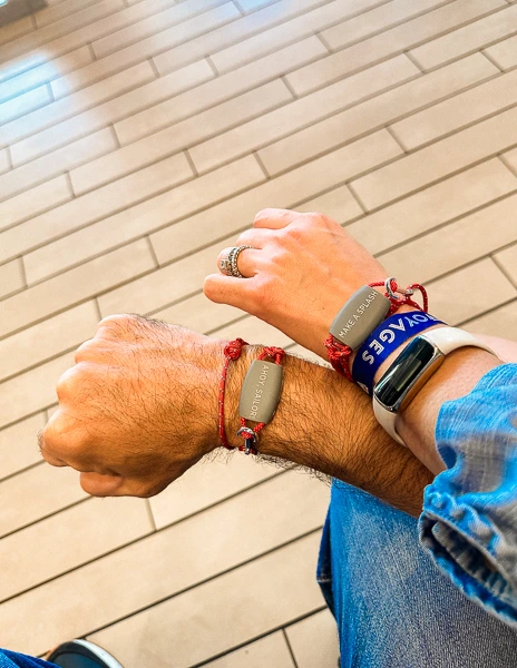 Two wrists close together, each wearing a red Virgin Voyages Band, against a tiled floor background. The bands replace traditional cruise cards for onboard transactions and cabin access.