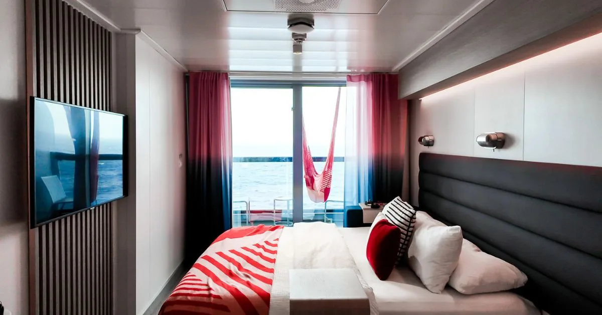 the virgin voyags scarlet lady xl sea cabin is a sleek design, featuring a large window with a sea view and hammock, a wall-mounted TV, and a comfortable bed with vibrant red and white bedding. The room is accented with colorful curtains