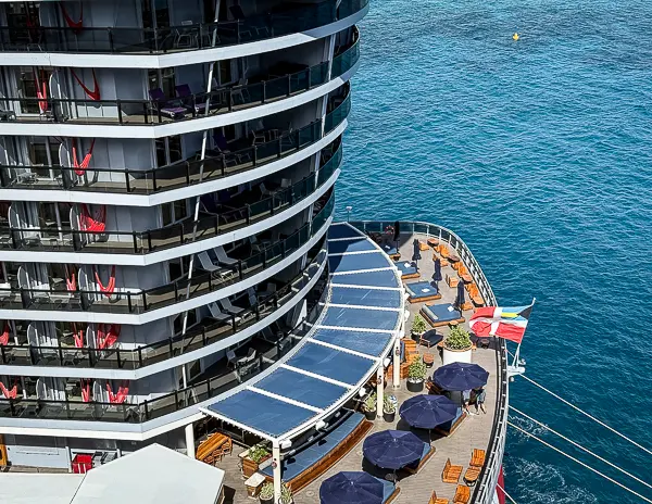 View of The Dock lounge at the aft of a Virgin Voyages ship with balcony cabins, red hammocks, umbrellas, and ocean backdrop.