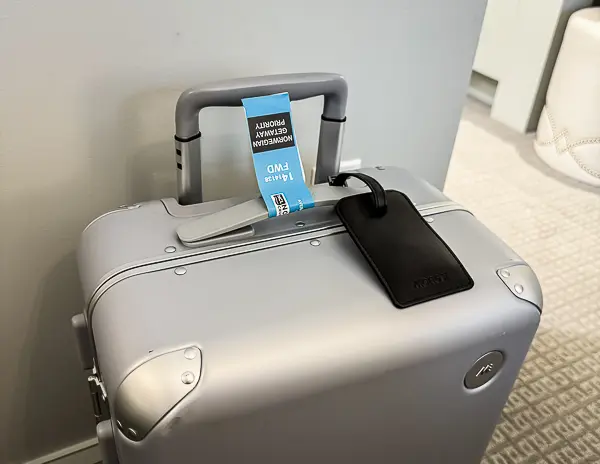 A silver suitcase with a blue "Norwegian Getaway Haven Priority" luggage tag attached to the handle.