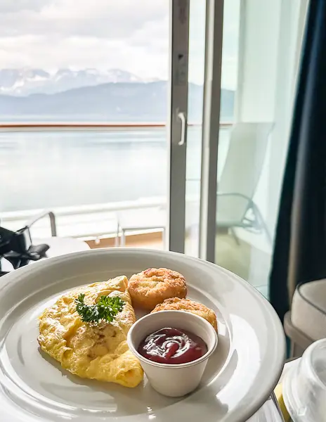Plate with an omelet, hash browns, and ketchup cup served as room service on a Norwegian Cruise Line ship, with a balcony view of snow-capped mountains in Alaska.