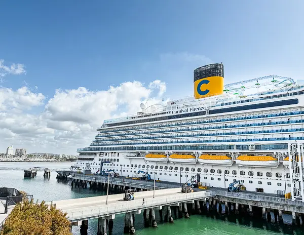 Carnival Firenze docked at the Long Beach Cruise Terminal, showing the Costa-style yellow funnel with a large blue “C” against a partly cloudy sky.