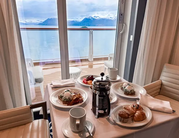 Table set for breakfast inside a Haven suite on Norwegian Encore with views of Glacier Bay and snow-capped mountains through the balcony doors
