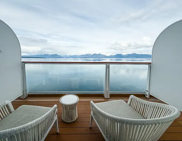 Spacious balcony with two lounge chairs and a table overlooking the water from a Penthouse Suite in The Haven on Norwegian Encore, featuring an upgraded larger balcony