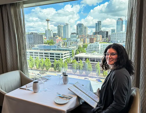 Woman sitting at a window table in The Haven Restaurant aboard Norwegian Encore with a view of downtown Seattle and the Space Needle in the background