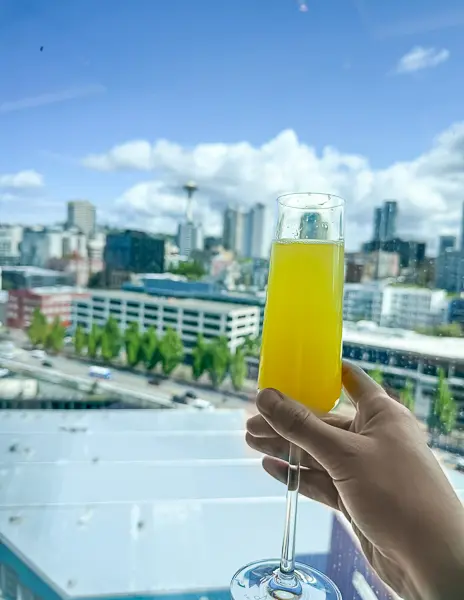 Hand holding a mimosa in front of a large window overlooking downtown Seattle and the Space Needle from The Haven Restaurant aboard Norwegian Encore