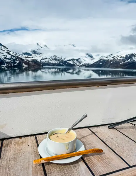 Cup of salmon chowder on a table aboard Norwegian Encore’s Haven Sundeck with snow-covered mountains and Glacier Bay in the background