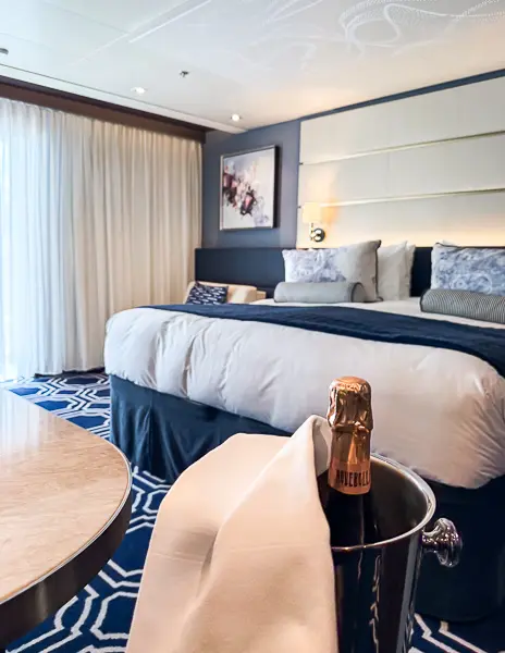Interior of The Haven Penthouse Suite on Norwegian Encore featuring a neatly made bed, balcony access, and a welcome bottle of sparkling wine on ice