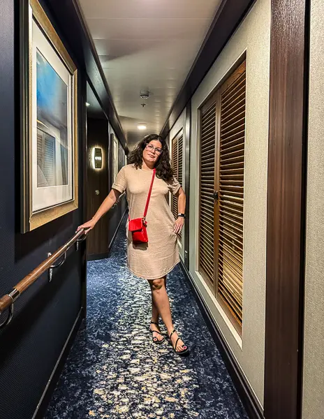 Woman (kathy) wearing a short-sleeve tan dress with black sandals and a red crossbody bag, standing in a cruise ship hallway before dinner on an Alaska cruise