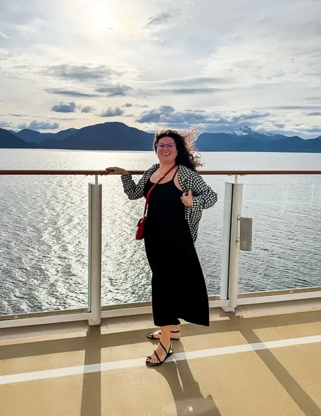 Woman (kathy) wearing a black midi dress by Pact, printed cardigan, and sandals with a red crossbody bag, standing on a cruise ship deck with ocean and mountains in the background during an Alaska cruise