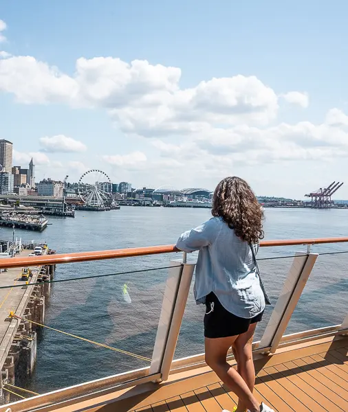Woman (kathy) facing the Seattle skyline from a cruise ship deck, wearing black shorts and a light blue button-down shirt, with the Seattle Great Wheel visible in the background